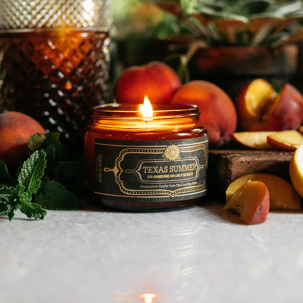 Texas Summer Candle burning surrounded by fresh peaches, mint leaves, and a textured amber glass, evoking a warm and inviting outdoor ambiance.