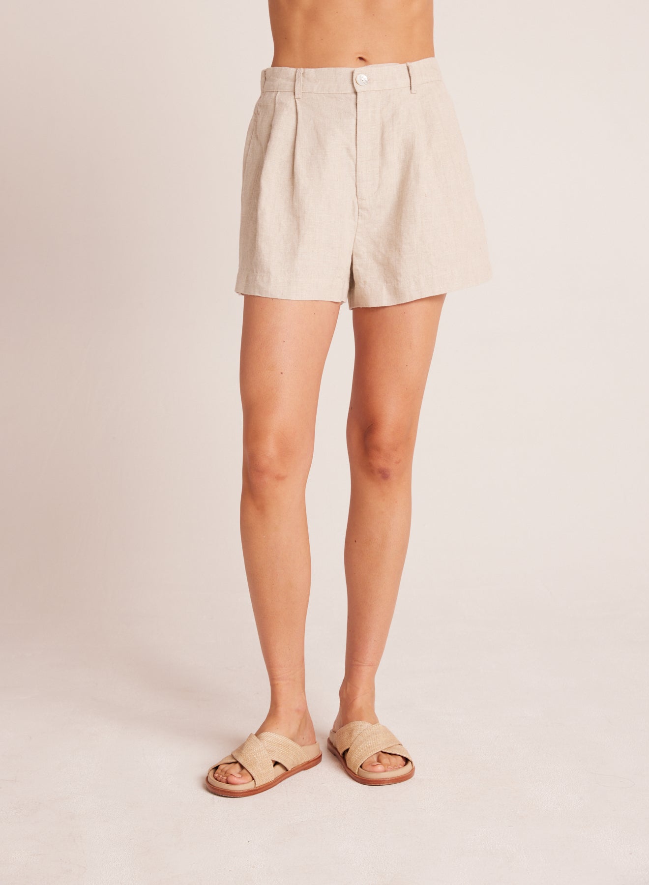 A front view of a person wearing light beige Pleat Front Trouser Short paired with matching slip-on sandals, showcasing a relaxed yet polished summer style.