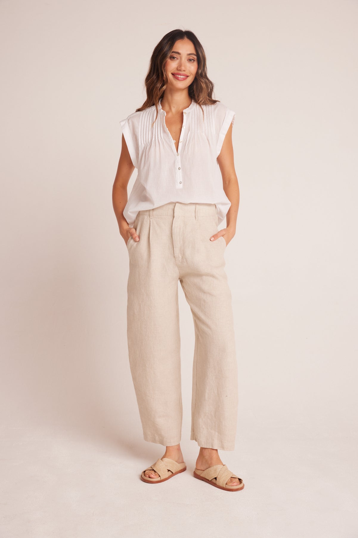 Woman wearing a relaxed fit, light beige Relaxed Pleat Front Trouser paired with a white sleeveless pleated blouse and casual tan sandals, standing against a plain background.