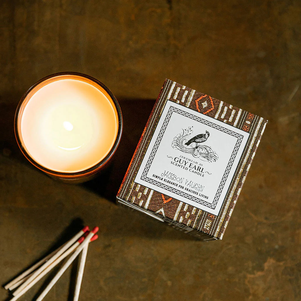Lit Guy Earl Candle beside its decorative box with a rustic pattern, accompanied by several wooden matches on a warm-toned surface, evoking a cozy atmosphere.
