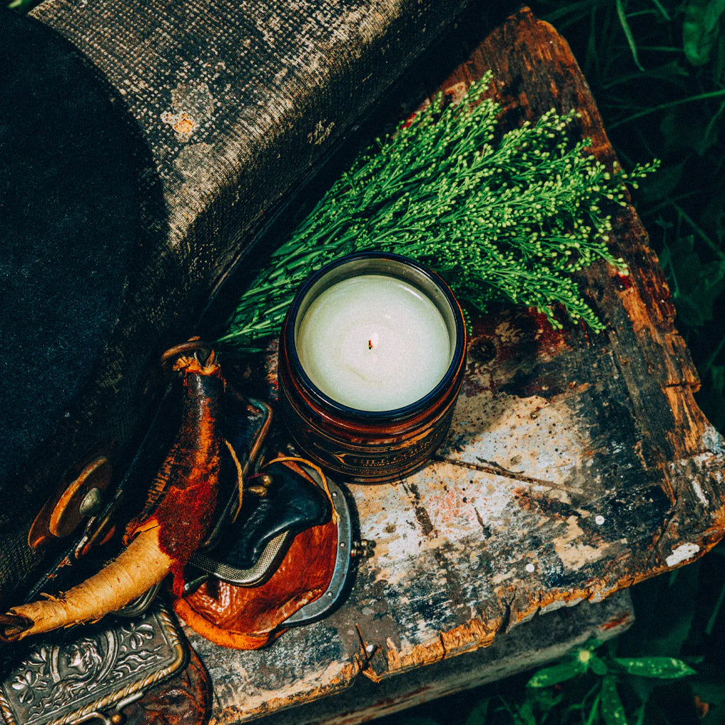 Hill Country Candle 16 Oz in a rustic amber glass jar surrounded by greenery and vintage leather and metal accessories, evoking authentic Hill Country style.