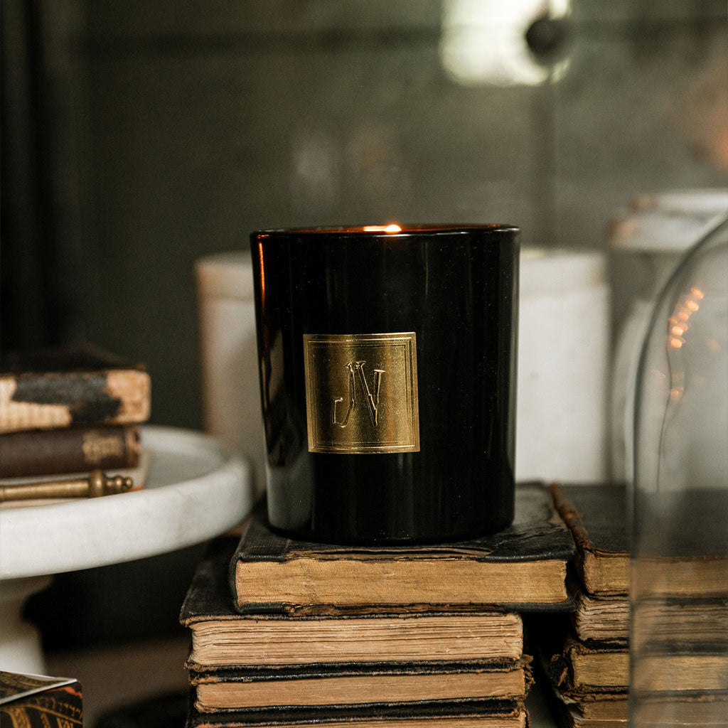Hyde Candle in a sleek black glass holder with a gold emblem, elegantly placed on vintage stacked books, evoking a warm, high-quality home fragrance atmosphere.
