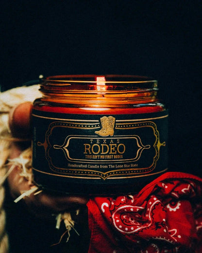 Close-up of a lit Texas Rodeo Candle 16 Oz in a dark amber jar with a western-style label featuring a cowboy boot, held by a hand next to a red bandana, evoking authentic Hill Country style.