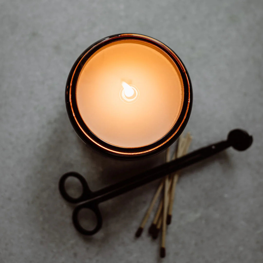 San Antonio Candle 16 Oz lit, glowing warmly in a dark container, accompanied by wooden matches and a black metal wick trimmer on a light textured surface.