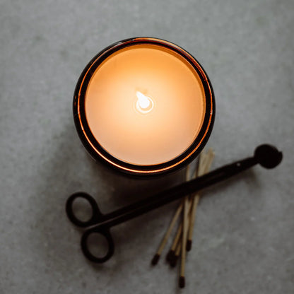 San Antonio Candle 16 Oz lit, glowing warmly in a dark container, accompanied by wooden matches and a black metal wick trimmer on a light textured surface.