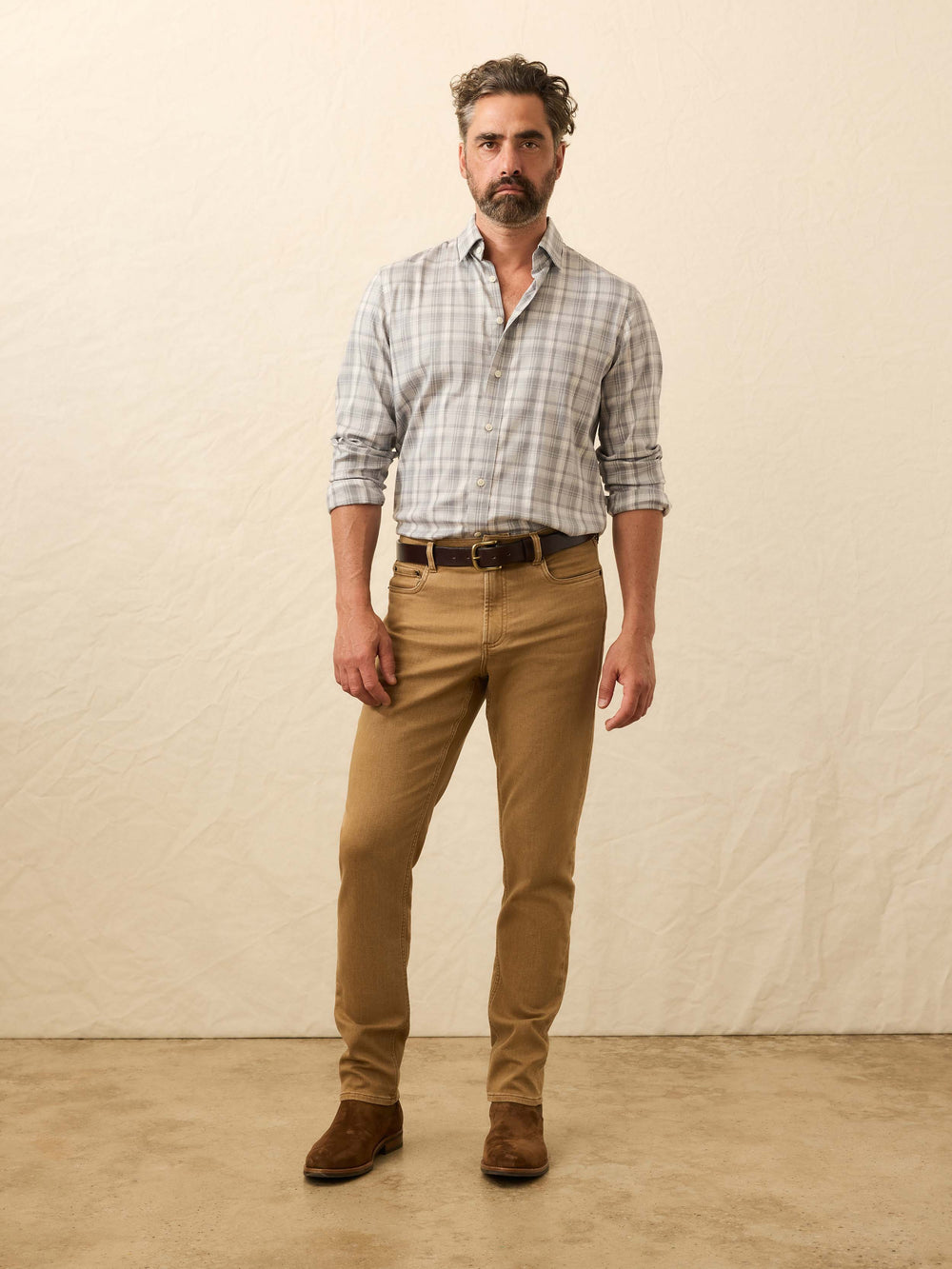 Man wearing a plaid shirt, brown trousers, and leather boots against a neutral backdrop