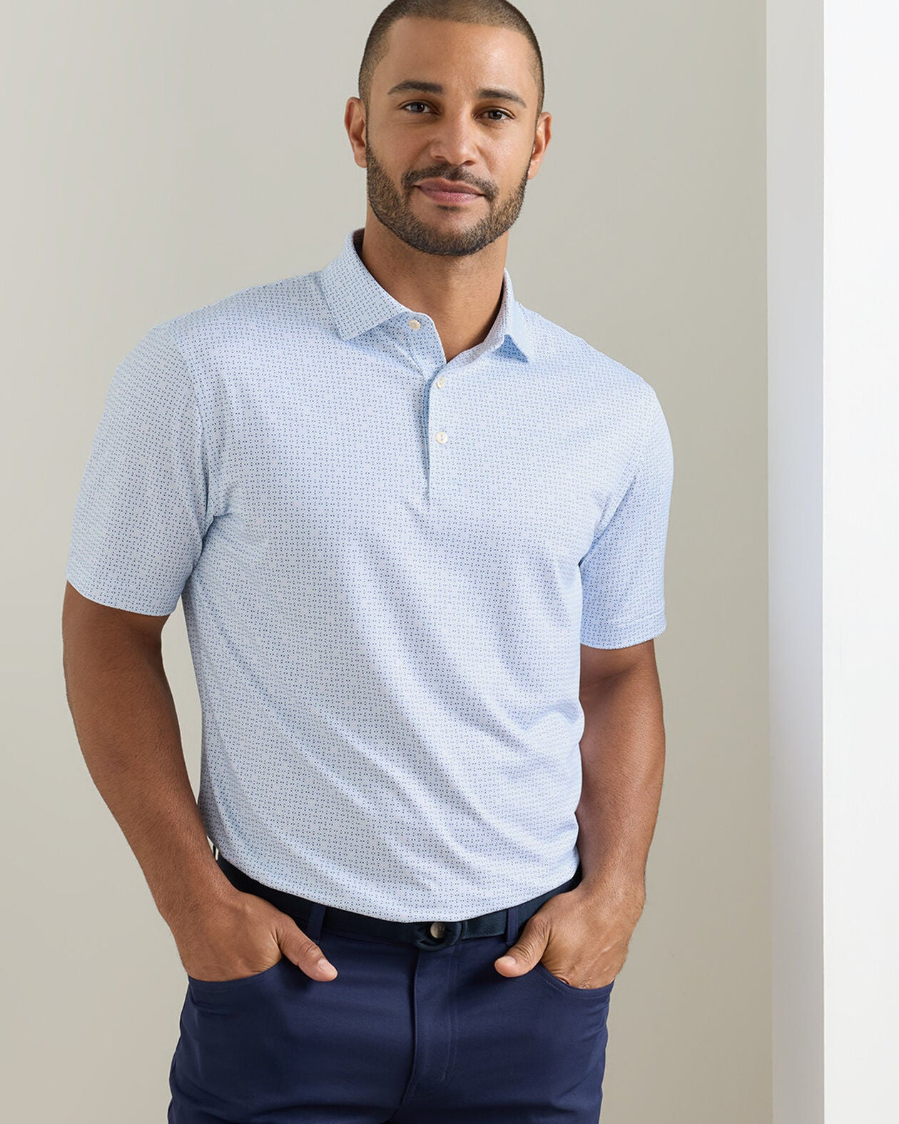 Man wearing a light blue patterned polo shirt and navy pants against a beige background