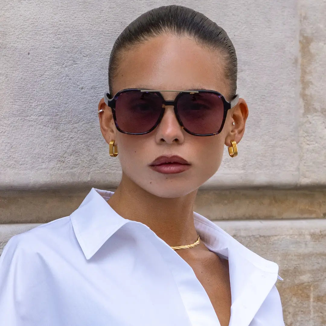 Woman wearing large black framed Spencer sunglasses with gradient lenses, gold hoop earrings, and a white collared shirt against a stone wall background.