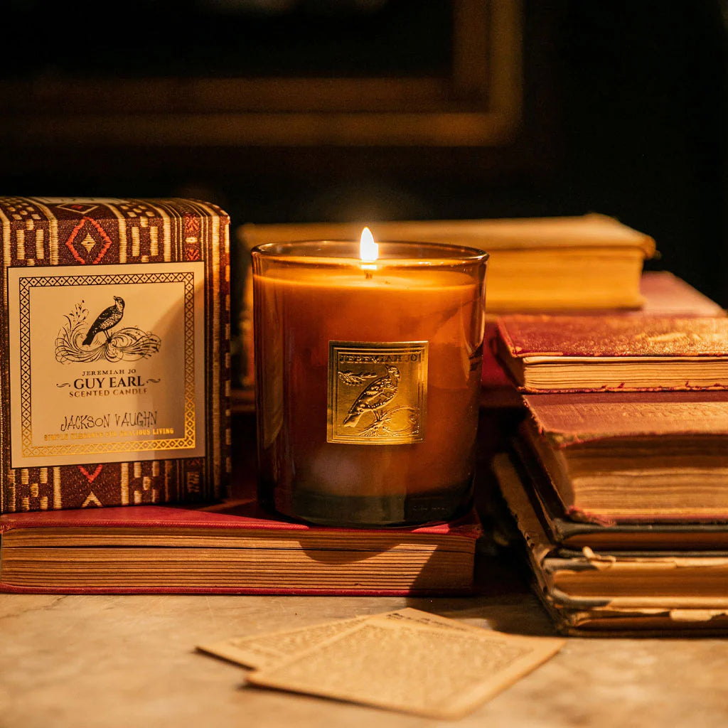 Guy Earl Candle lit beside its patterned box and stacked vintage books on a softly lit surface, evoking a warm, inviting home fragrance atmosphere.