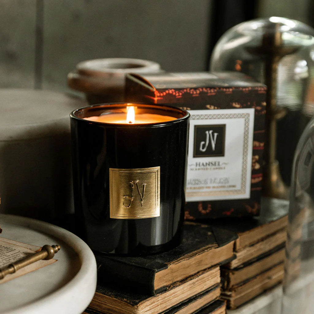 Lit Hansel Candle in a sleek black glass holder with a gold emblem, placed beside its decorative box on stacked vintage books, creating a warm, inviting ambiance.