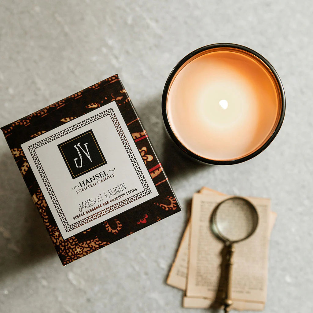 Hansel Candle in a decorative box next to a lit candle in a glass holder and a vintage magnifying glass on aged paper, showcasing a high-quality home fragrance product.
