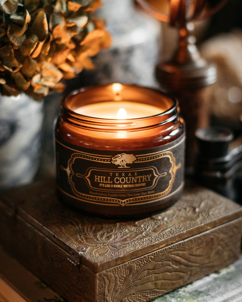 Hill Country Candle 16 Oz in a brown glass jar, lit and placed on a decorative wooden box, showcasing warm, rustic ambiance and premium home fragrance from Fredericksburg, Texas.