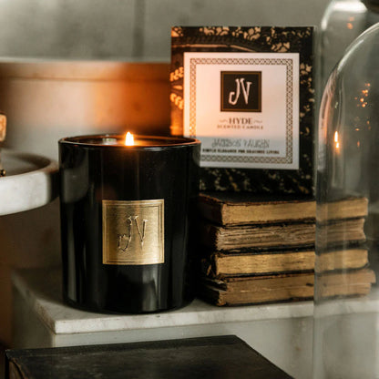 Lit Hyde Candle in a black glass container with a gold emblem, displayed beside vintage books and its decorative packaging, reflecting a warm, elegant ambiance.