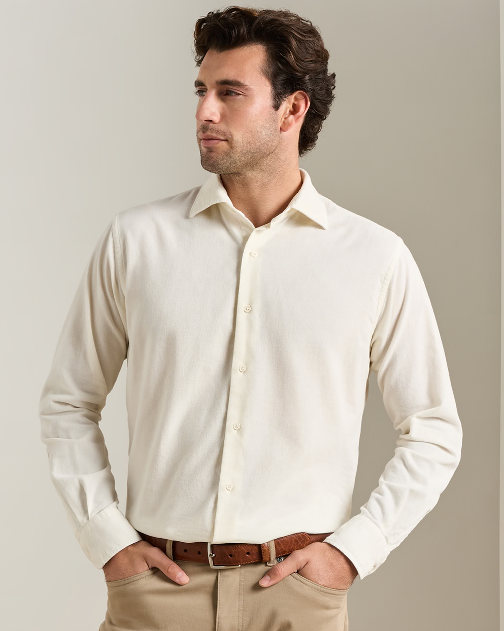 A man wearing a light cream-colored long-sleeve button-down shirt, khaki pants, and a brown leather belt, standing against a neutral beige background.