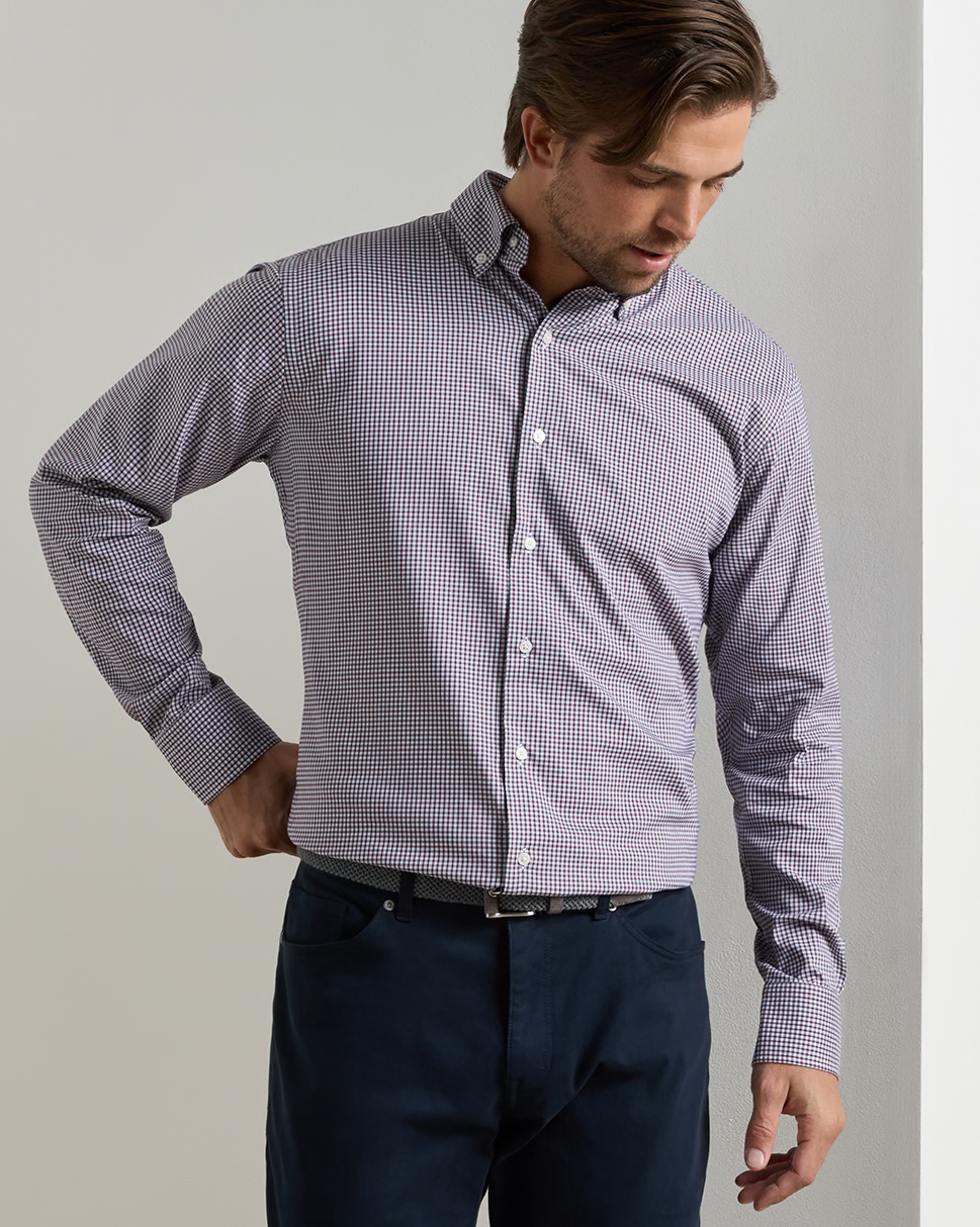 A man wearing a light purple and white micro-checkered long-sleeve button-down shirt with dark navy trousers, standing against a neutral light gray background.
