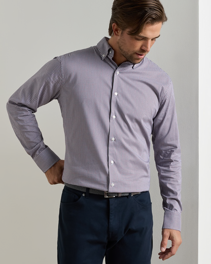 A man wearing a light purple and white micro-checkered long-sleeve button-down shirt with dark navy trousers, standing against a neutral light gray background.