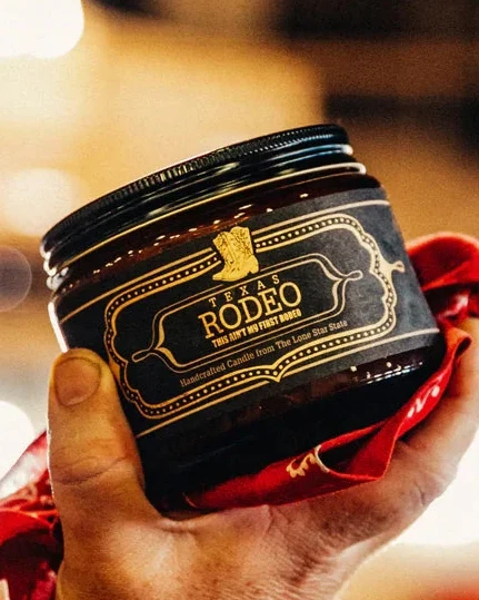 Hand holding Texas Rodeo Candle 16 Oz in a dark jar with a black lid, wrapped with a red bandana, showcasing handcrafted home fragrance inspired by authentic Hill Country style.