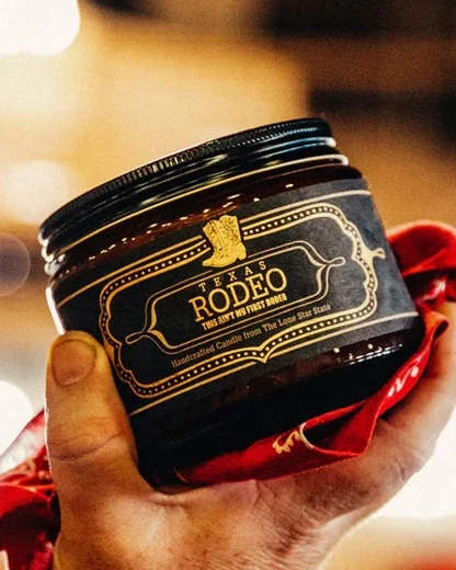 Hand holding Texas Rodeo Candle 16 Oz in a dark jar with a black lid, wrapped with a red bandana, showcasing handcrafted home fragrance inspired by authentic Hill Country style.