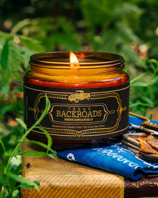 Candle labeled 'Texas Backroads' on a wooden block with greenery in the background