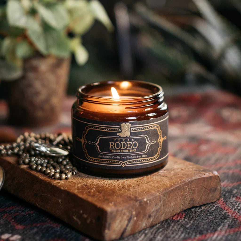 Texas Rodeo Candle 16 Oz burning in a brown glass jar on a wooden board, styled with rustic decor reflecting authentic Hill Country style.