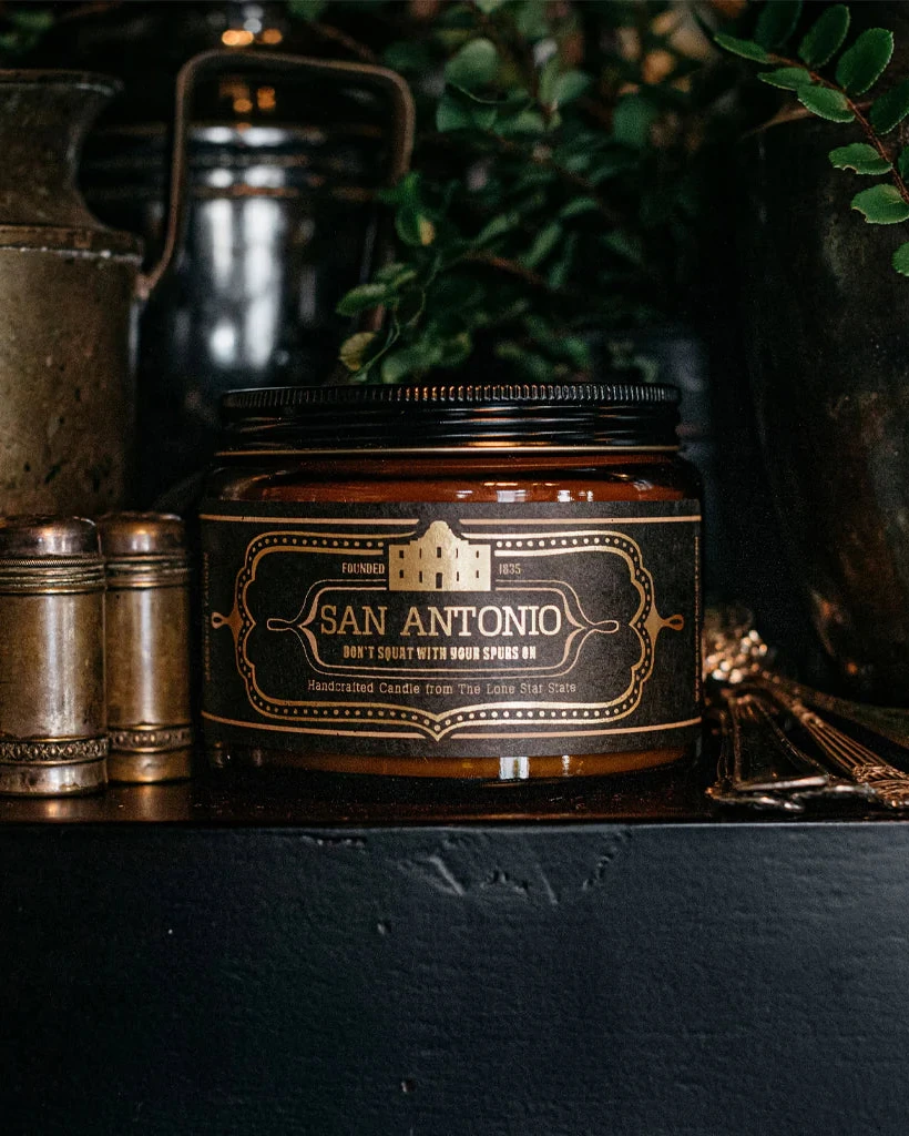 San Antonio Candle 16 Oz in a brown glass jar with a vintage-style label, surrounded by rustic decor and greenery, embodying premium boutique home fragrance.