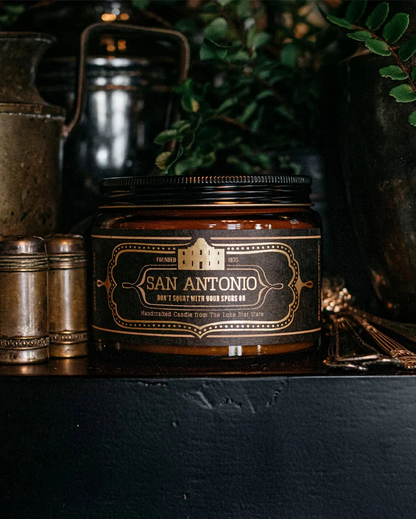 San Antonio Candle 16 Oz in a brown glass jar with a vintage-style label, surrounded by rustic decor and greenery, embodying premium boutique home fragrance.