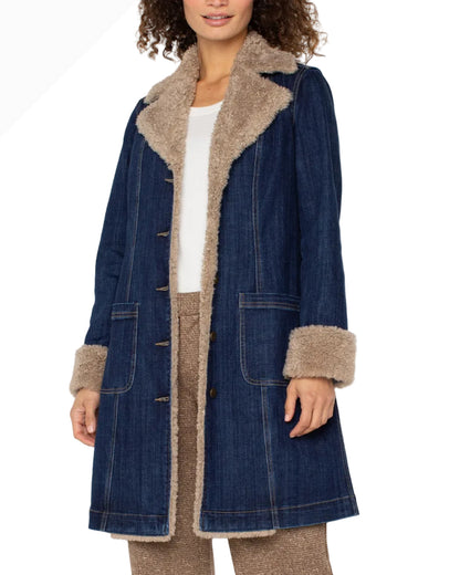 A woman wearing a dark blue denim-style coat with a beige sherpa lining on the collar, cuffs, and front opening, styled over a white top and brown pants against a plain white background.